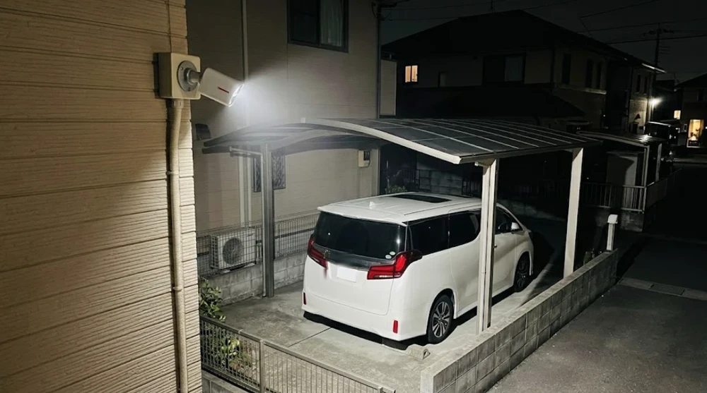夜の一戸建ての駐車場。外壁の防犯カメラのライトが、停めてある白いミニバンを照らしている