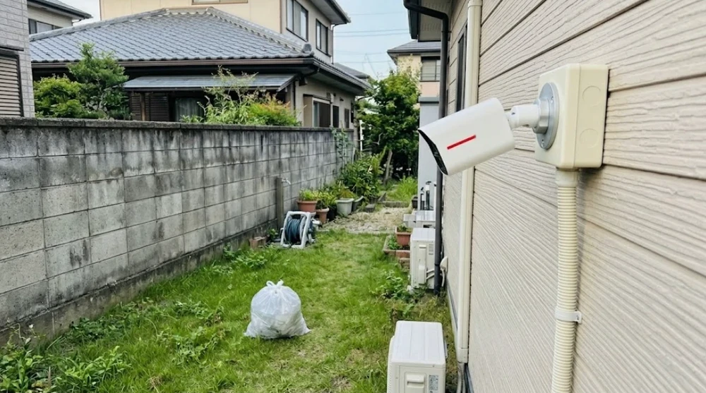 ブロック塀に面した庭に投げ込まれたゴミ袋と、それを見下ろす外壁の防犯カメラ